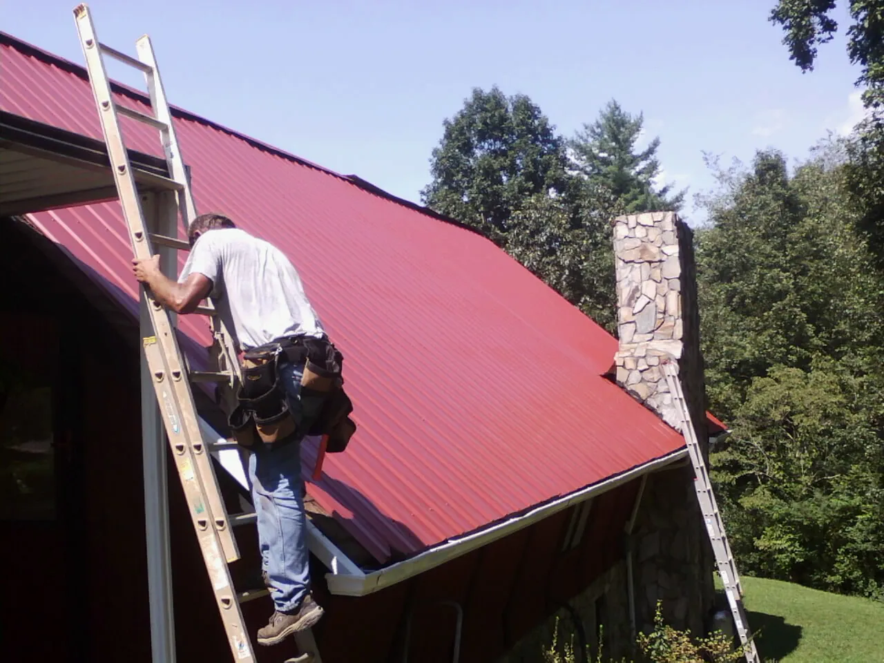 Roofer on a ladder beside a red metal roof and stone chimney — Soffit & Fascia Repair by Roofing Brothers in Convent